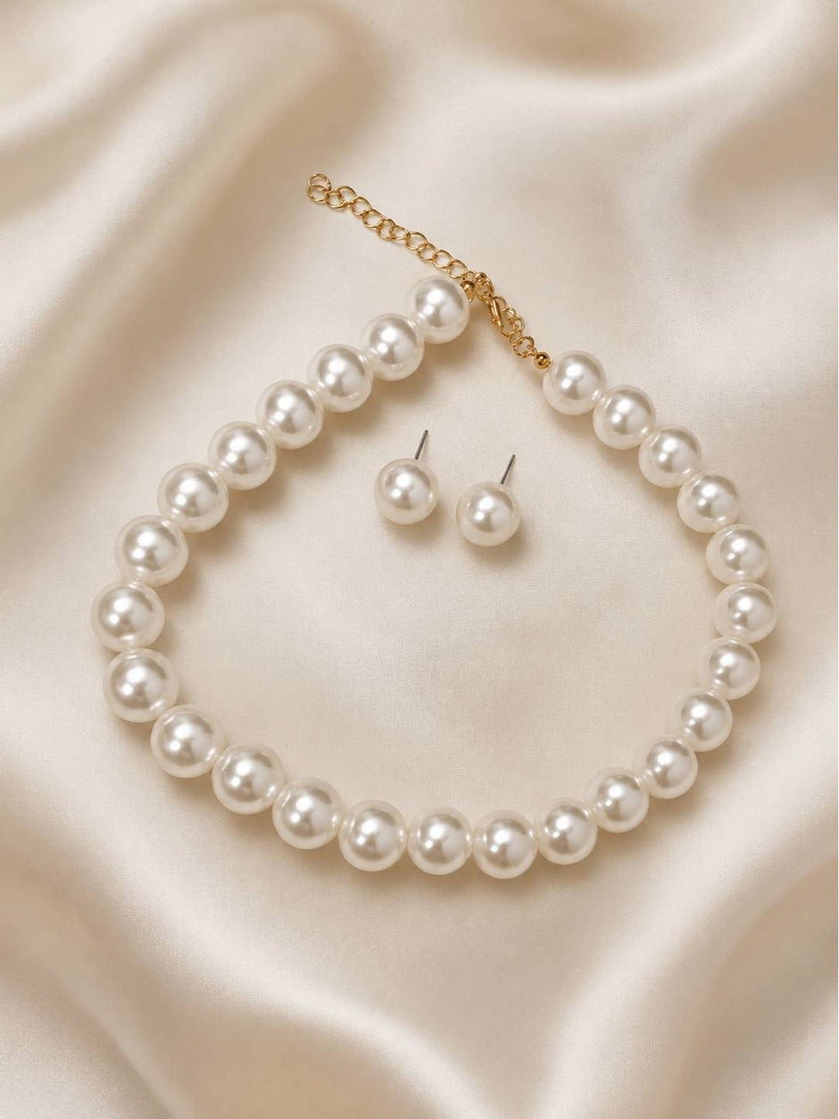 White pearl choker necklace laid flat on cream satin fabric beside a pair of pearl stud earrings with gold extender chain visible