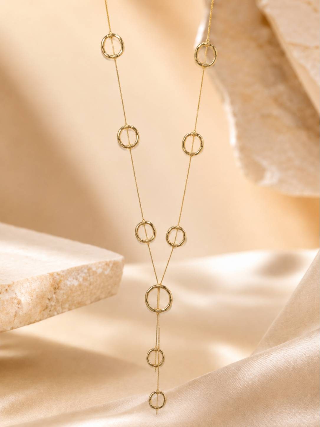 Gold lariat necklace with hammered open-circle charms styled on a warm stone and silk fabric backdrop