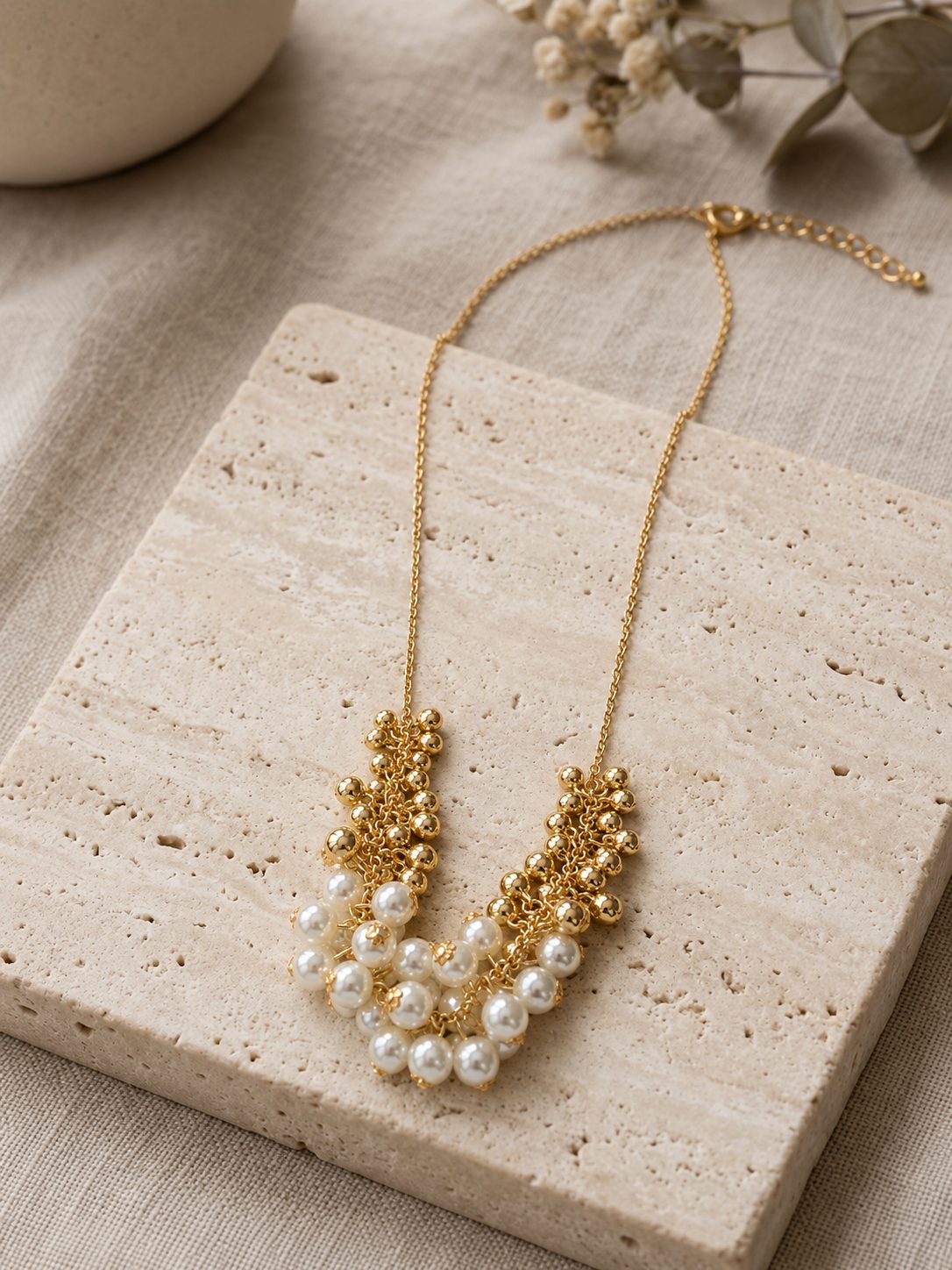 Pearl and gold bead cluster necklace displayed on a travertine stone tile with eucalyptus sprigs in background