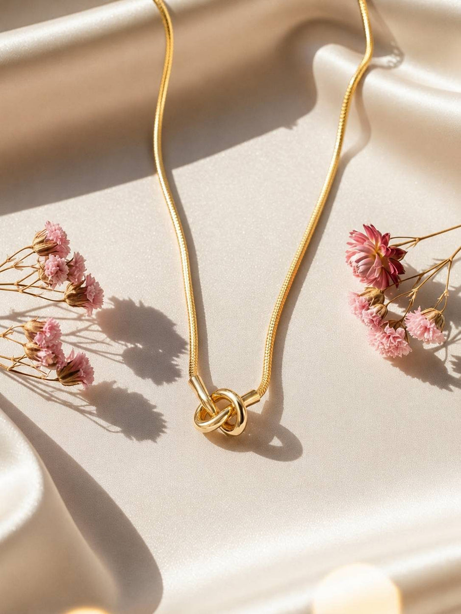 Gold-plated knot pendant necklace laid flat on cream satin surrounded by dried pink flowers
