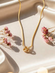 Gold-plated knot pendant necklace laid flat on cream satin surrounded by dried pink flowers