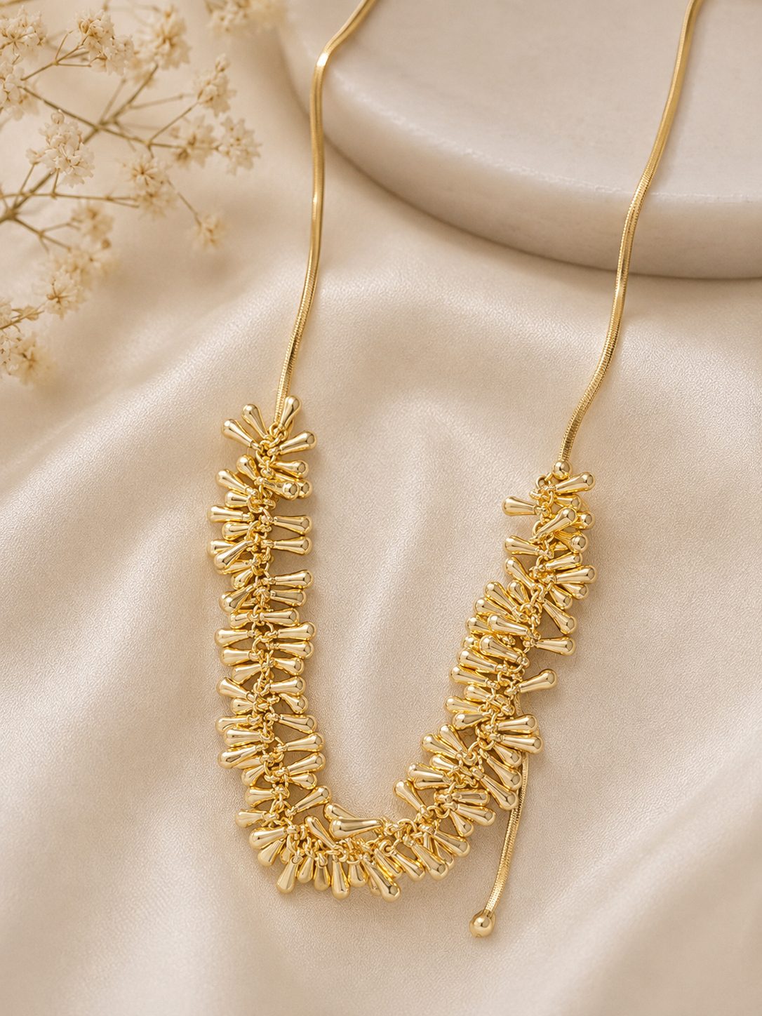 Gold-plated spike drop necklace laid flat on cream satin fabric beside dried flowers
