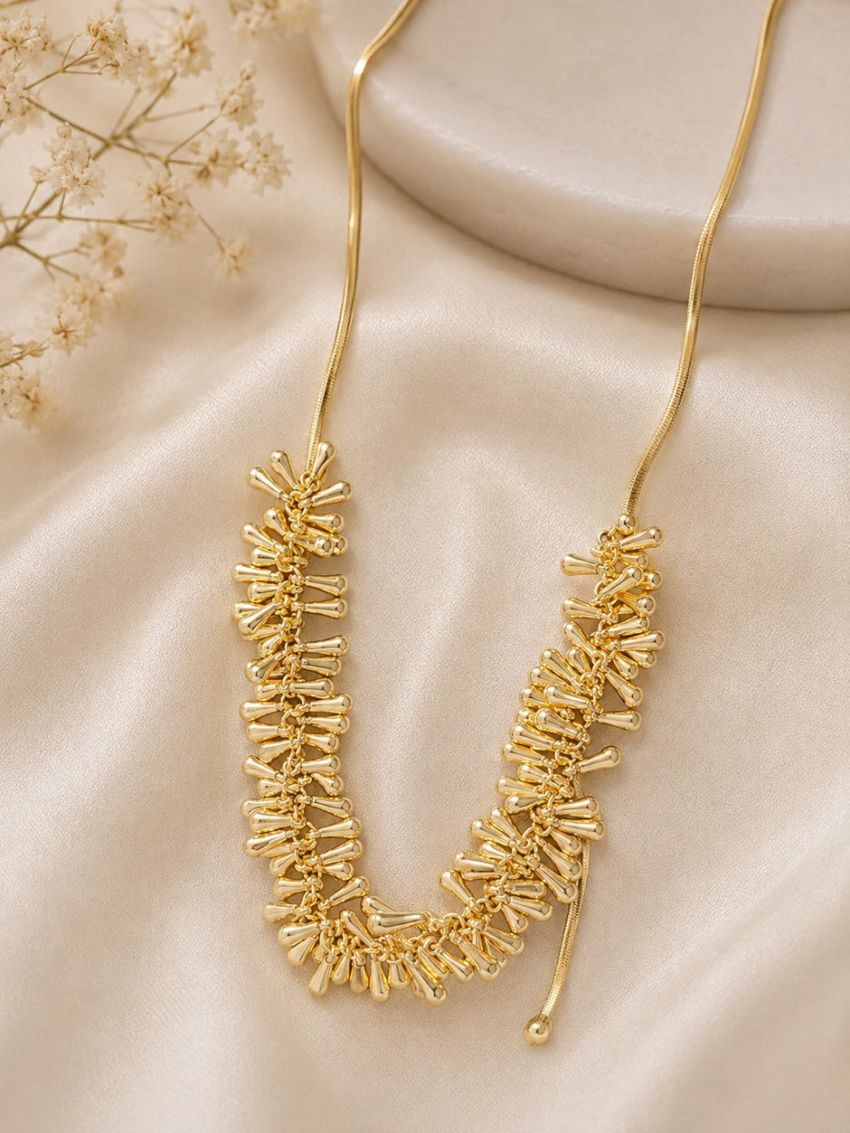 Gold-plated spike drop necklace laid flat on cream satin fabric beside dried flowers