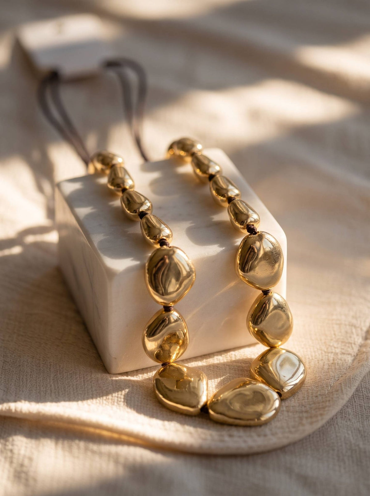 Chunky gold pebble necklace draped over a white ceramic block in warm sunlight