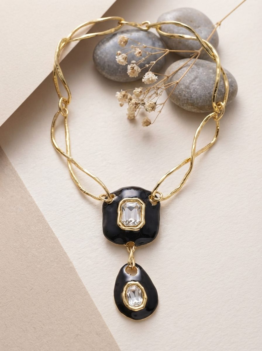 Black enamel crystal pendant necklace displayed overhead on beige paper with smooth pebbles and dried flowers