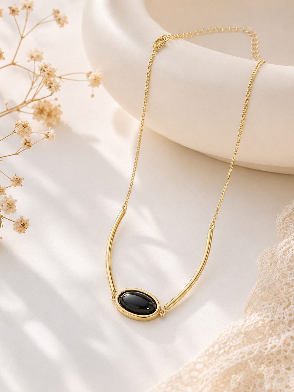 Gold-plated oval black stone pendant necklace laid flat on white fabric beside dried flowers