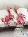 Pair of pink crab drop earrings with push-back closures resting on open pages of a book