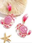 Two pink enamel crab earrings displayed near a white fan shell and a gold starfish on white background