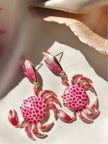 Pink enamel crab drop earrings with gold-tone finish resting on fabric beside a large seashell in sunlight