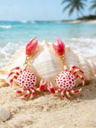 Flat lay of pink crab drop earrings on sandy beach surface with ocean and palm trees in background