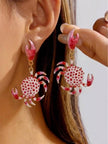 Pink and white dotted crab earrings propped inside a large conch shell on a sandy beach