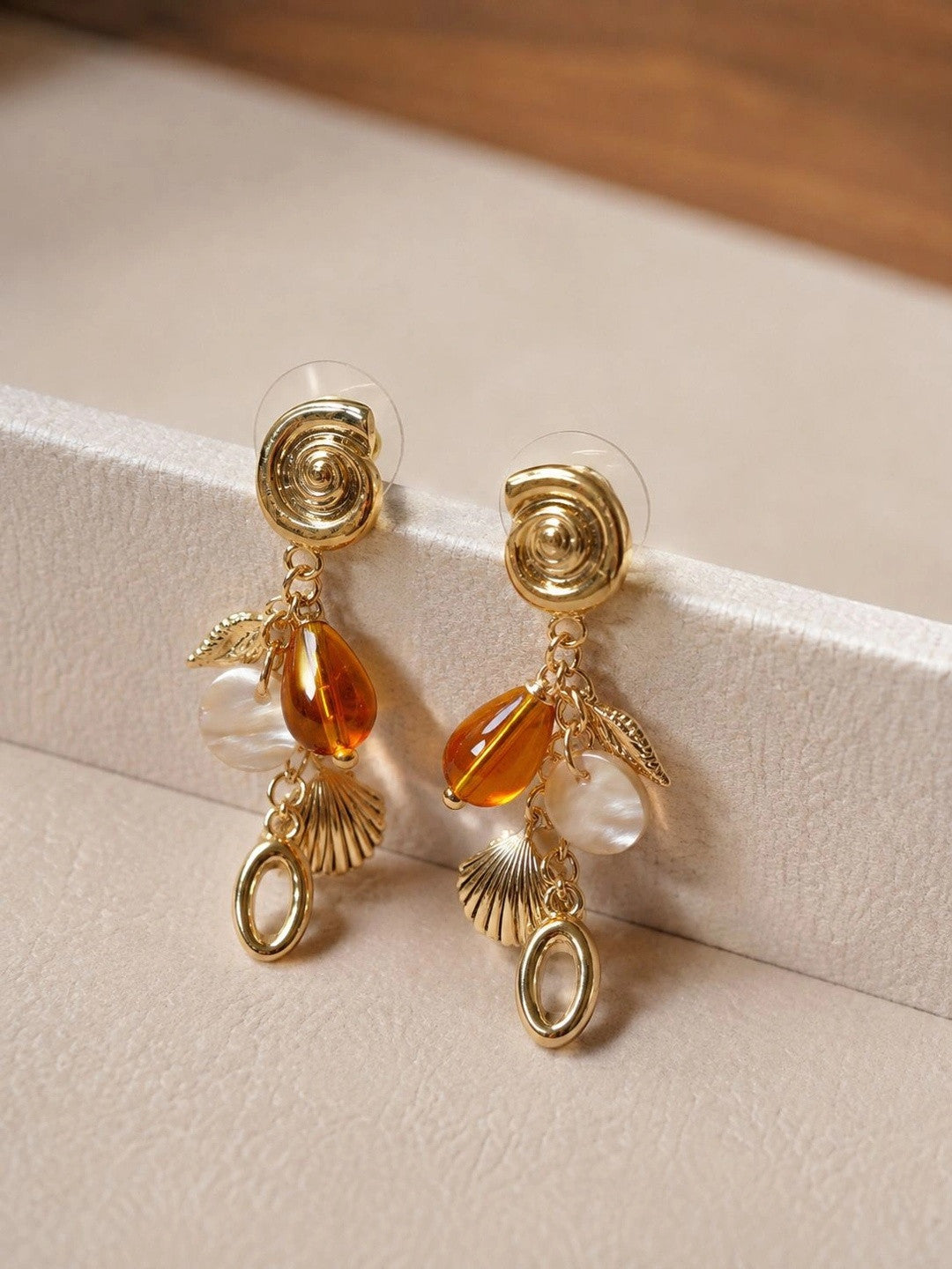 Pair of gold shell charm drop earrings with amber resin bead resting on a white jewellery box edge