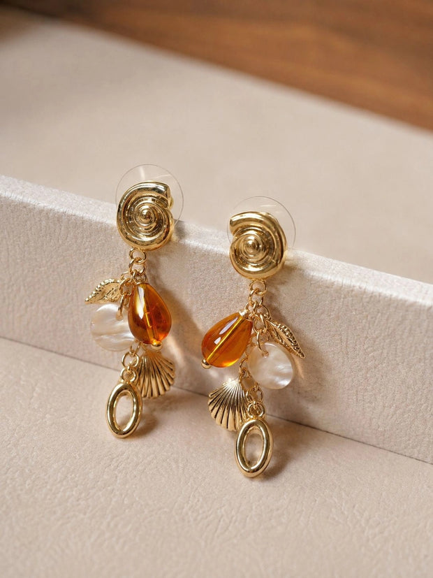 Pair of gold shell charm drop earrings with amber resin bead resting on a white jewellery box edge