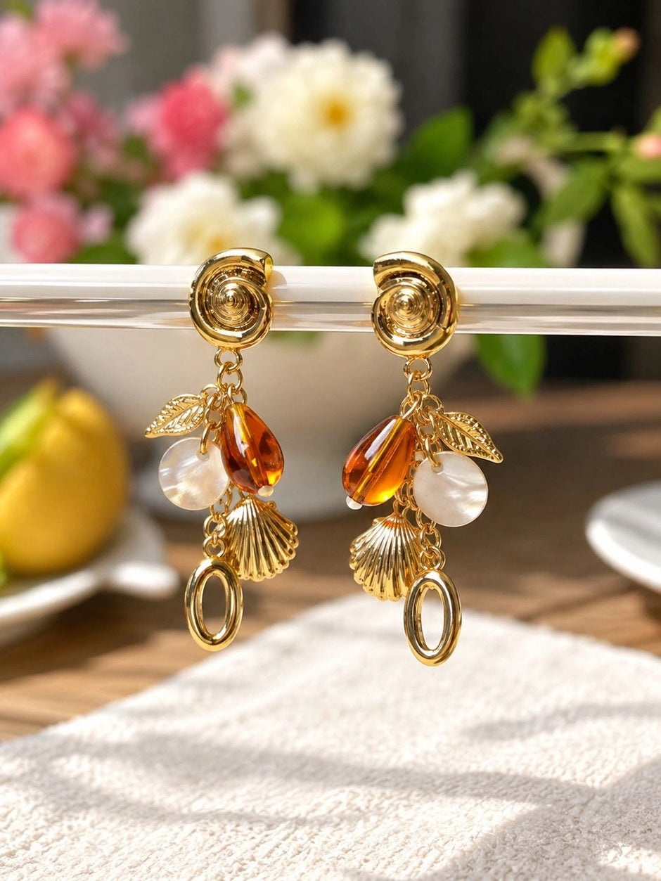 Gold shell charm drop earrings hanging from a glass rod with floral background