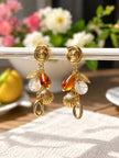 Gold shell charm drop earrings hanging from a glass rod with floral background