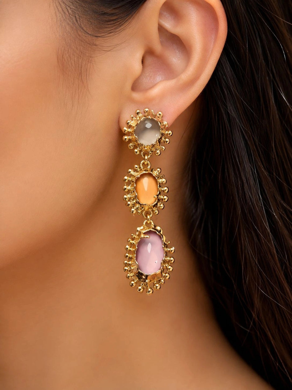 Close-up of gold-plated triple drop earring worn on ear, showing grey, orange and lilac oval cabochons in beaded frames