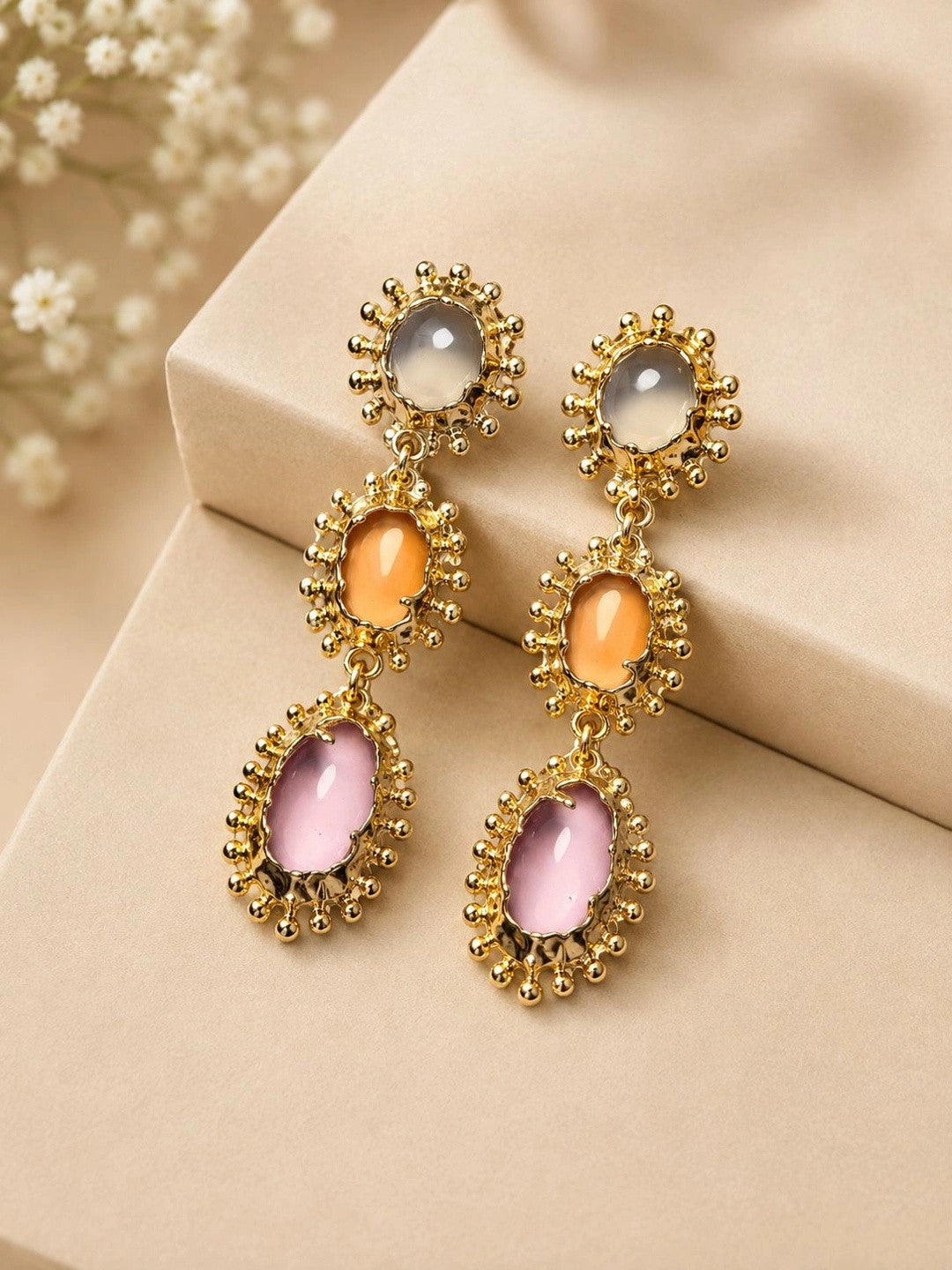 Pair of gold-plated triple drop earrings laid flat on cream surface with dried floral backdrop