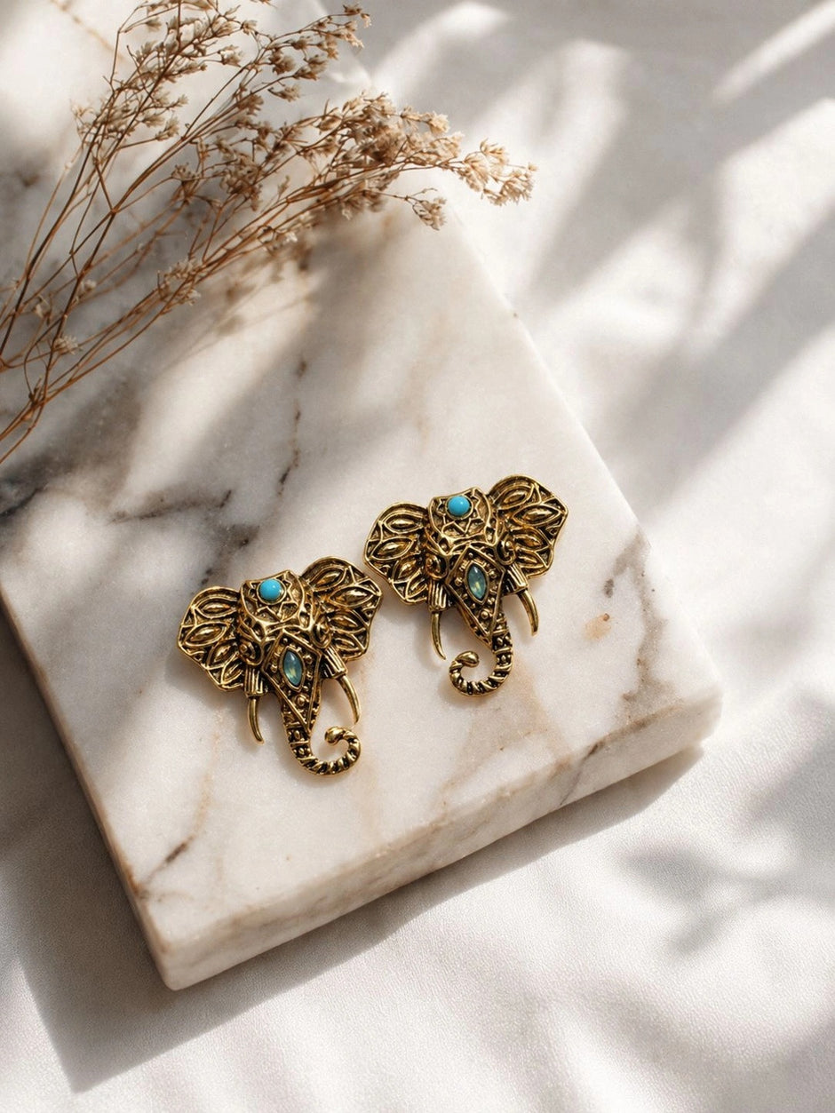 Two antique gold elephant stud earrings placed on white marble surface with dried floral stems in background