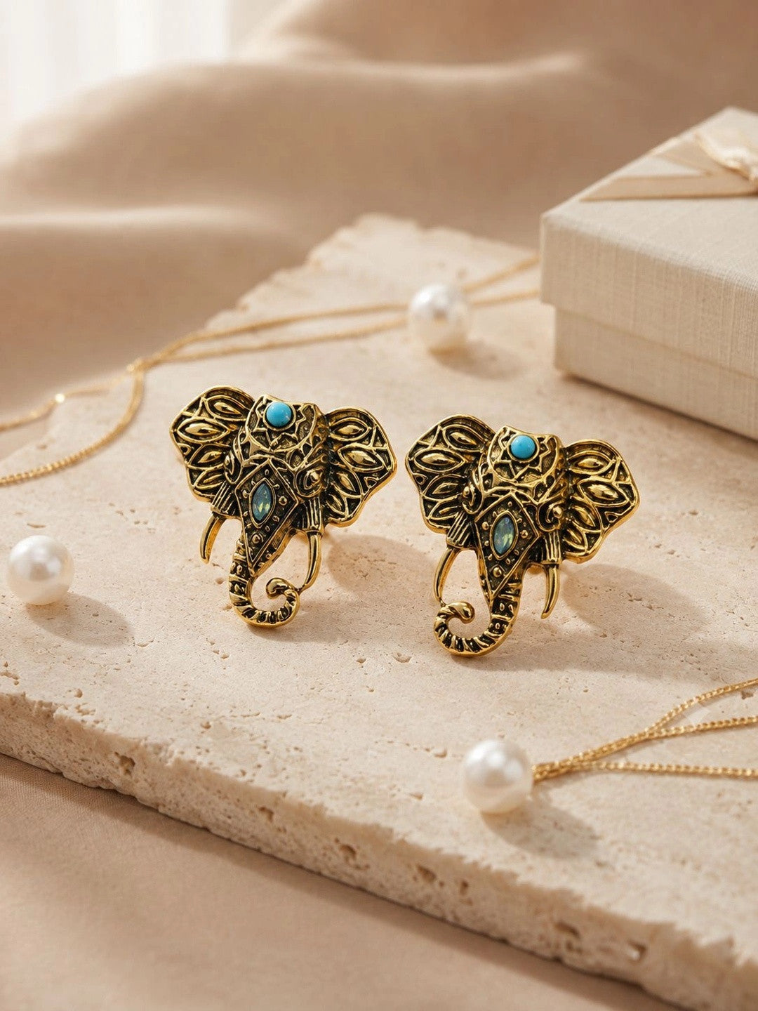 Antique gold elephant studs displayed on stone surface surrounded by pearls and gold chain styling props