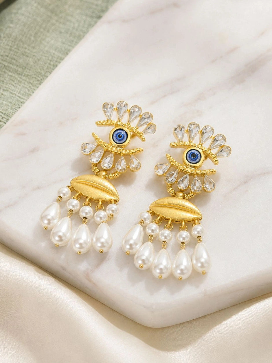 Pair of gold-plated evil eye drop earrings with crystals and pearl tassels on white marble