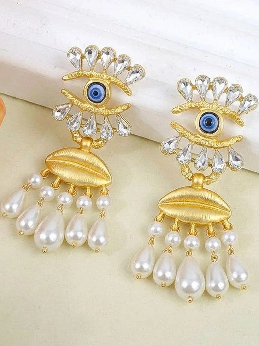 Close-up of gold evil eye crystal earrings with faux pearl fringe on cream background