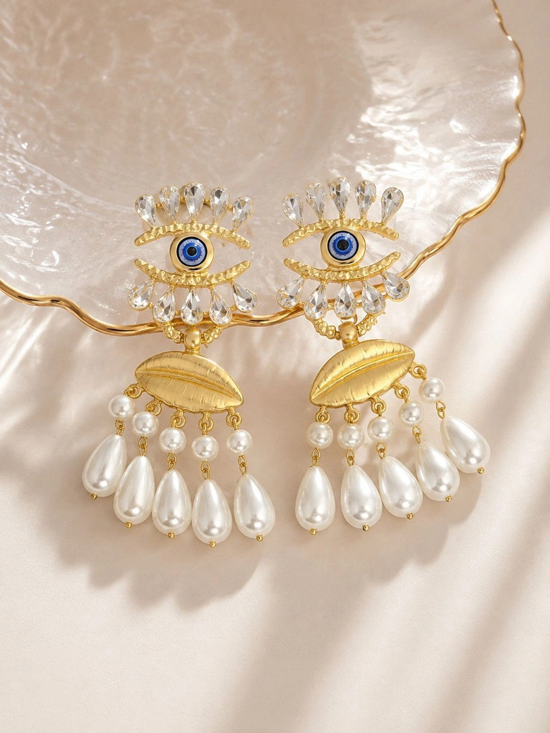 Both gold evil eye earrings displayed flat on a pearlescent shell dish in warm light