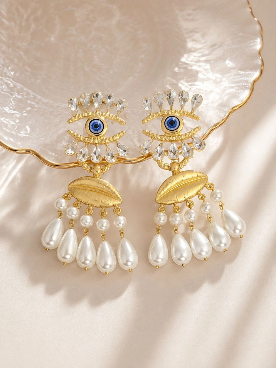 Both gold evil eye earrings displayed flat on a pearlescent shell dish in warm light