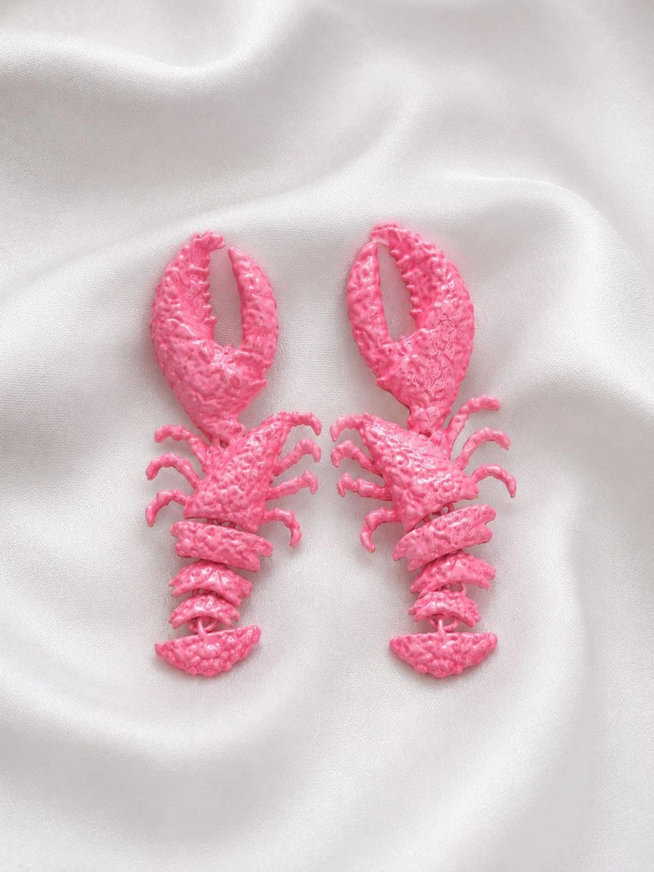 Pair of hot pink painted lobster drop earrings laid flat on white satin fabric