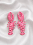 Pair of hot pink painted lobster drop earrings laid flat on white satin fabric