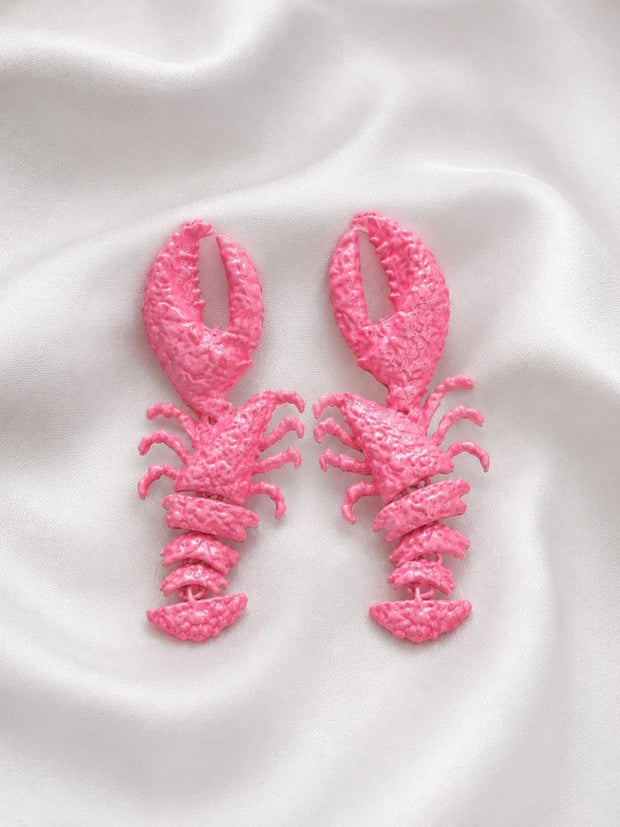 Pair of hot pink painted lobster drop earrings laid flat on white satin fabric