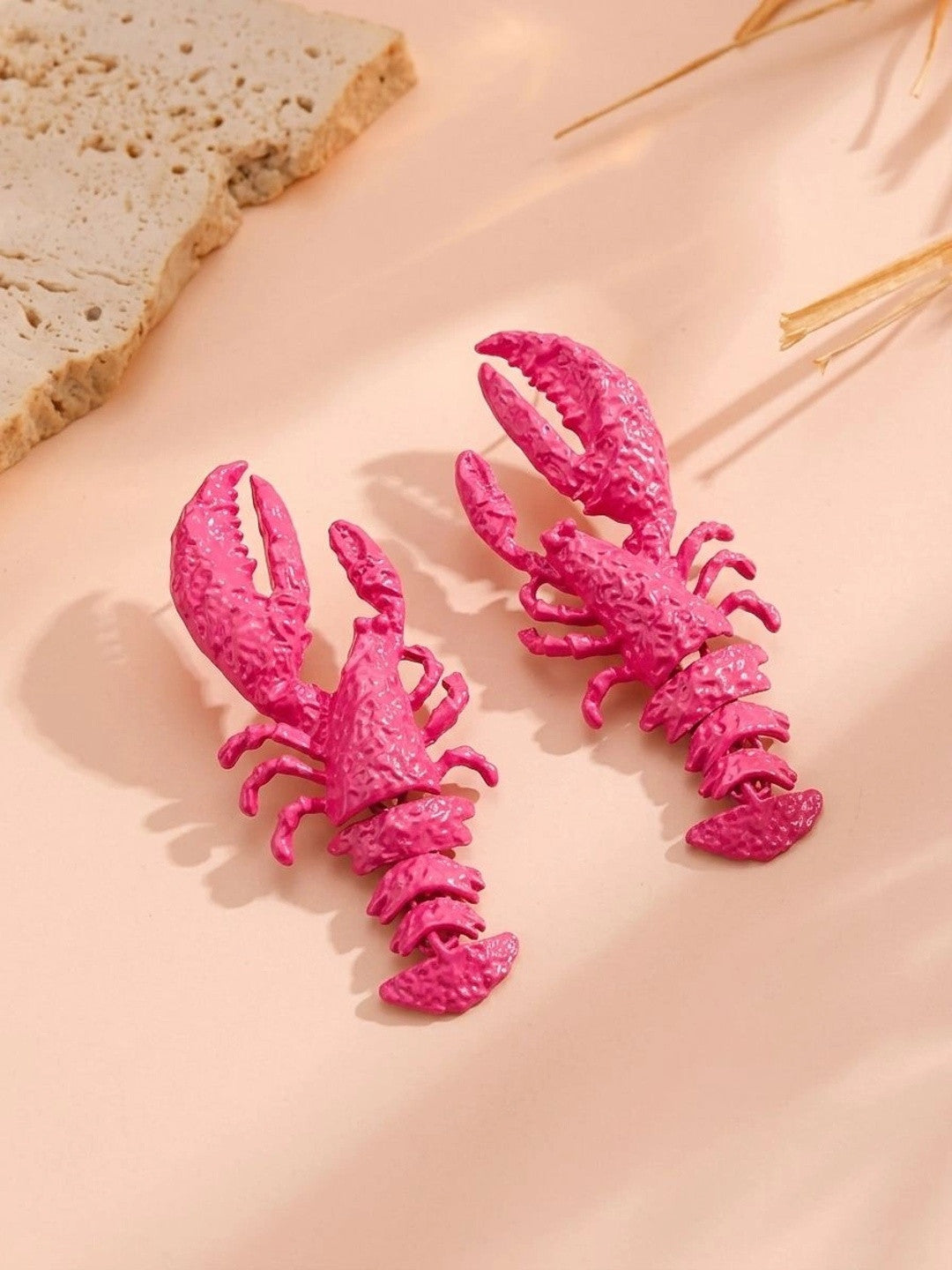 Pair of hot pink lobster earrings displayed on a peach surface with dried grass