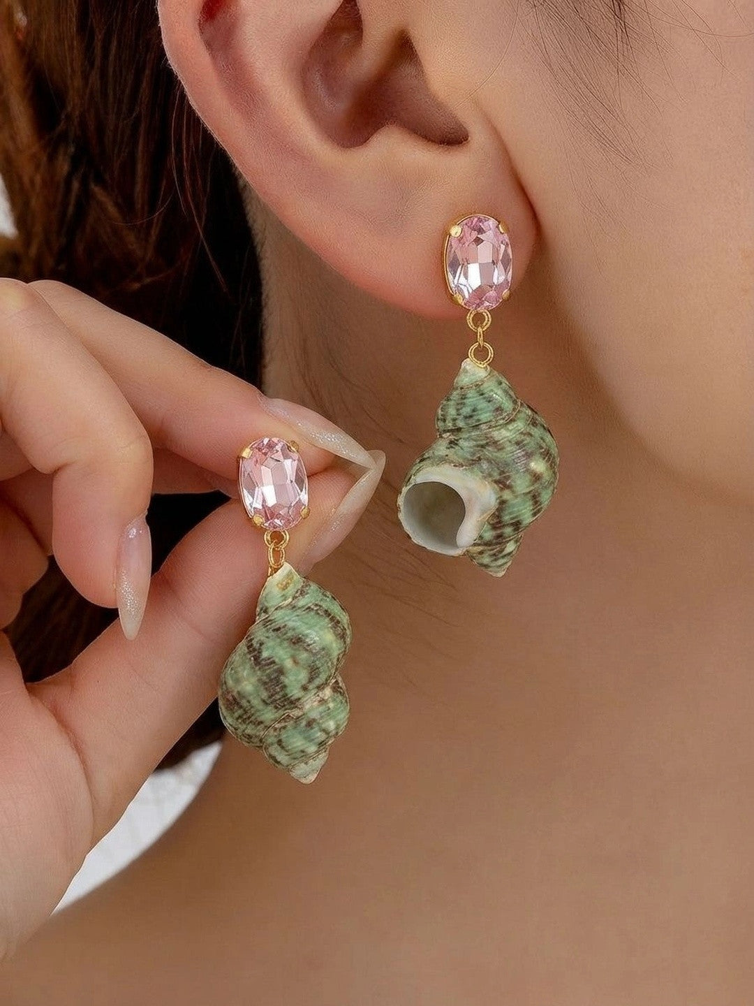 Woman wearing green turban shell drop earrings with pink crystal gold-toned studs, holding the second earring up for comparison