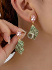 Woman wearing green turban shell drop earrings with pink crystal gold-toned studs, holding the second earring up for comparison