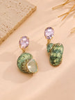 Green turban shell drop earrings with faceted pink crystal studs laid on a sandy beige surface beside dried grass stems