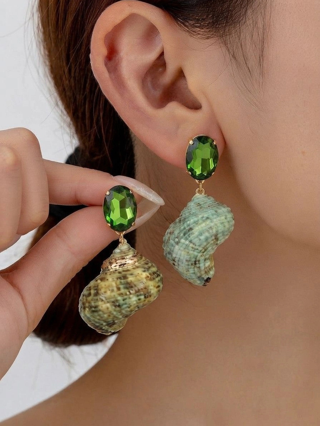 Woman wearing green crystal and natural seashell drop earrings, holding the second earring up for comparison