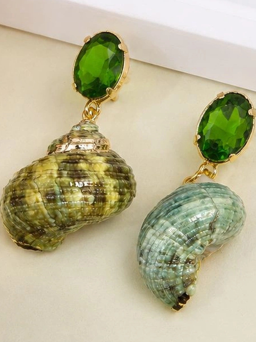 Pair of mismatched natural seashell drop earrings with faceted green crystal studs on a cream surface