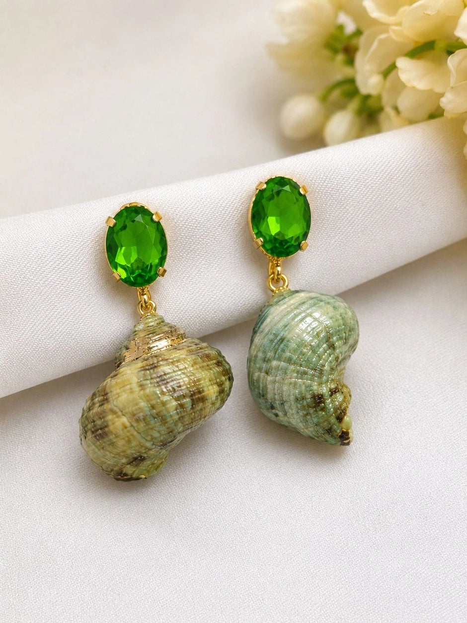 Two green crystal seashell drop earrings laid on white fabric beside small white flowers