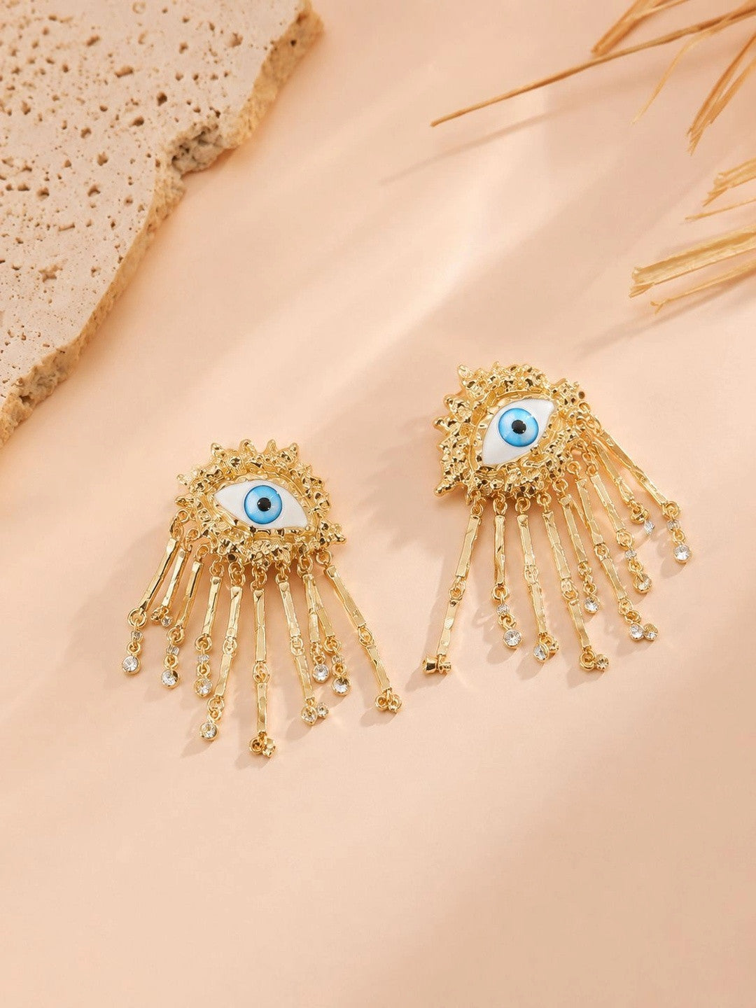 Pair of gold-plated evil eye tassel earrings with blue enamel and crystal tips on a peach surface