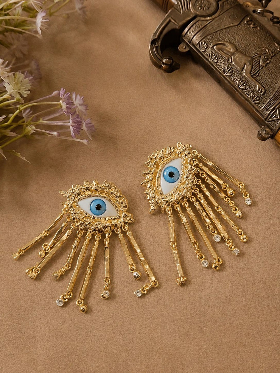 Two gold evil eye tassel earrings laid flat on a brown surface beside dried flowers and a vintage box