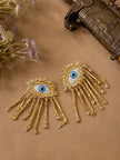 Two gold evil eye tassel earrings laid flat on a brown surface beside dried flowers and a vintage box