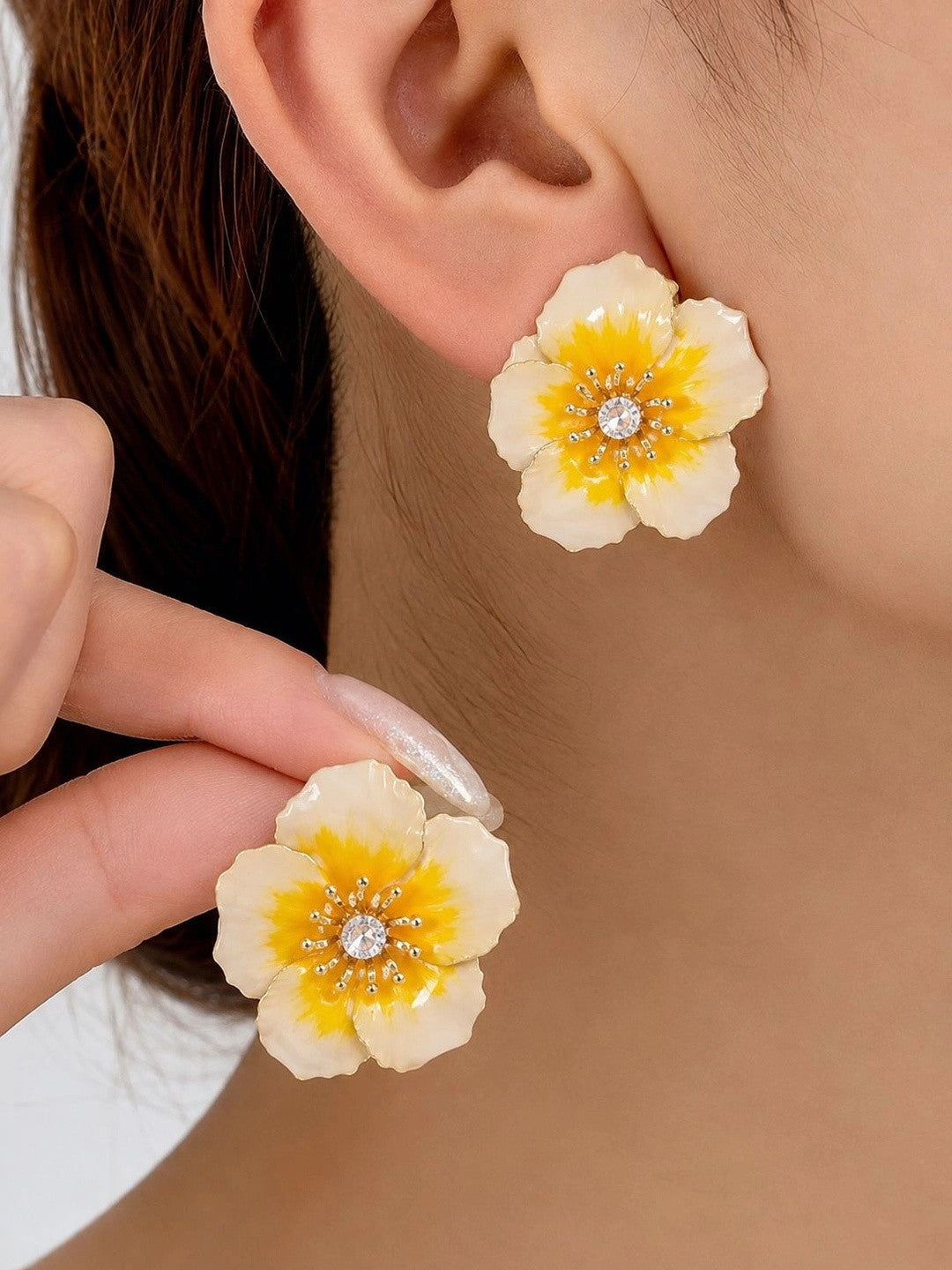 Woman wearing large ivory enamel floral stud earring, holding second earring close to ear