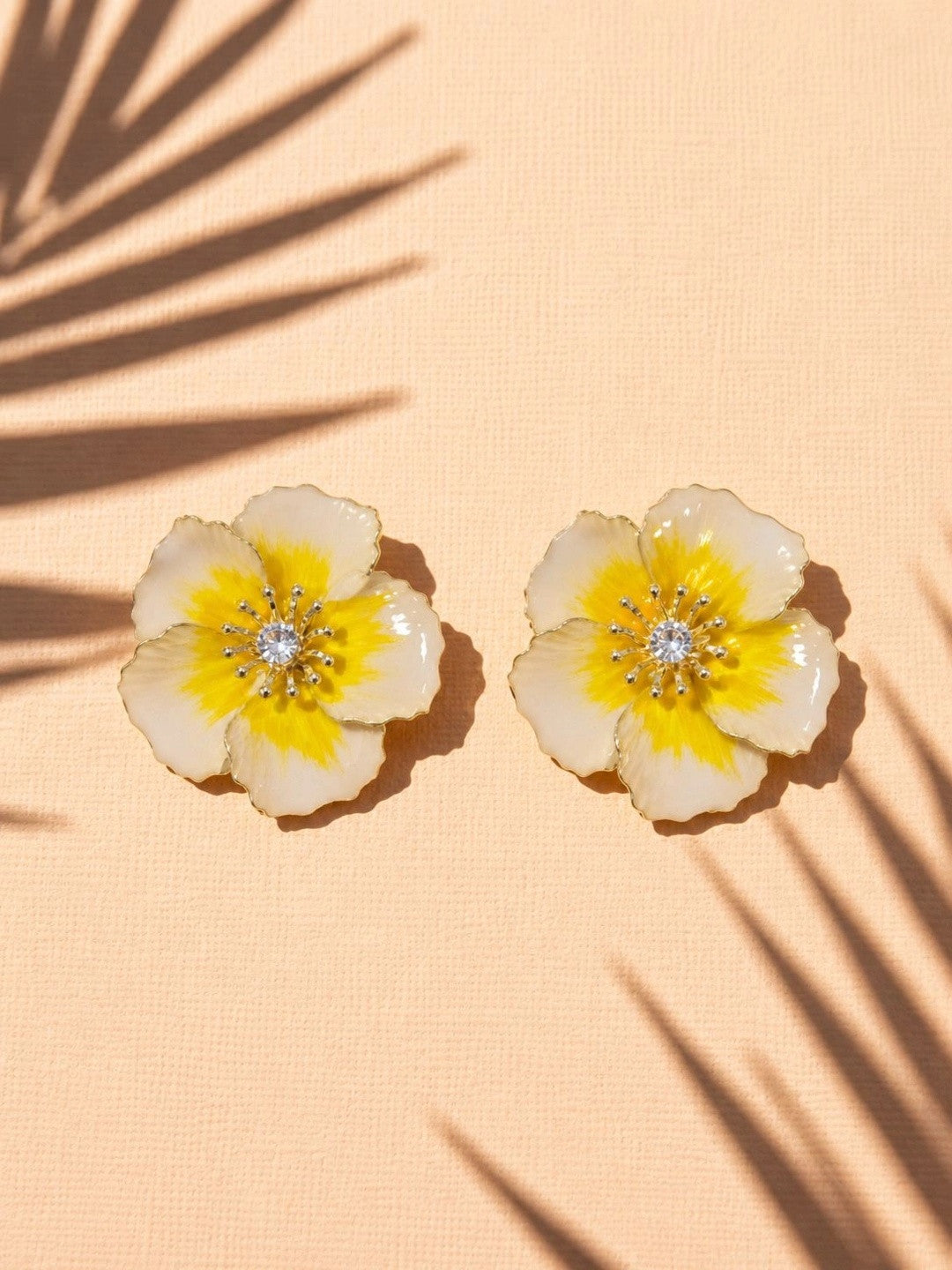 Two enamel floral stud earrings placed on a peach fabric surface with palm leaf shadows
