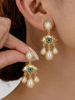 Gold-plated evil eye pearl drop earrings worn on a woman's ear, held between fingers with almond nails