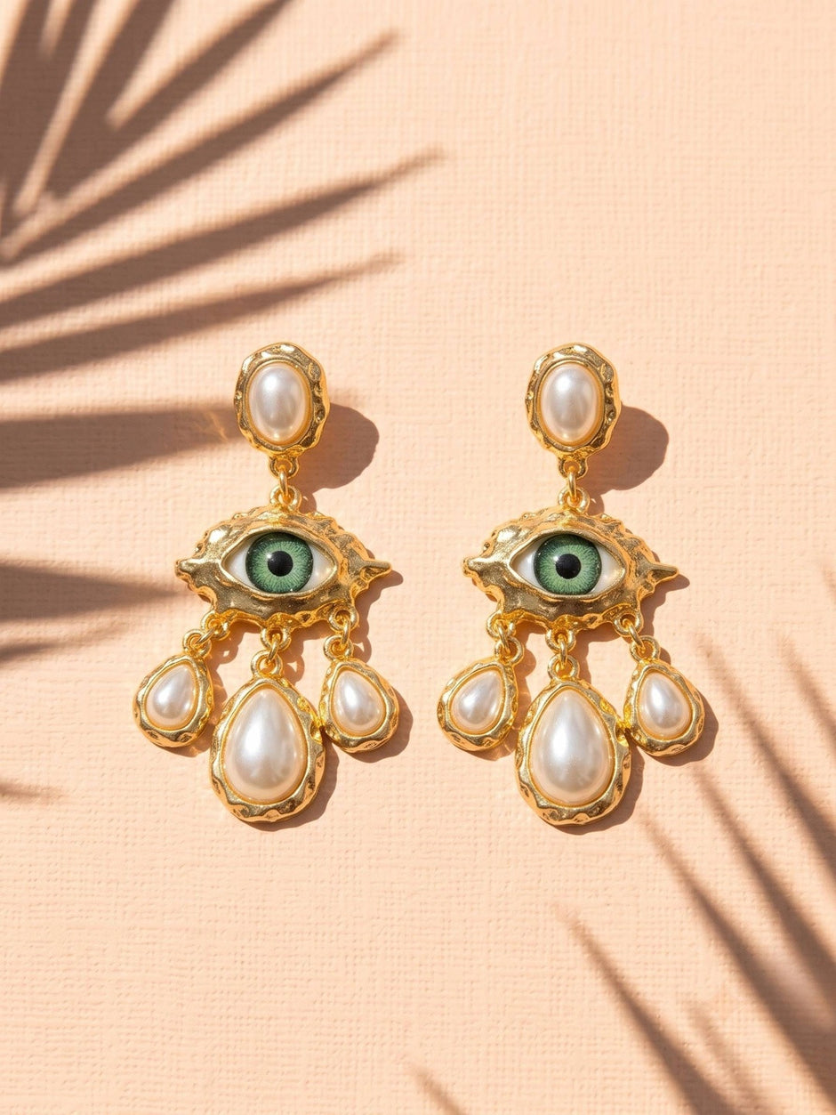 Pair of gold evil eye earrings with faux pearl drops laid flat on a peach surface with palm leaf shadow