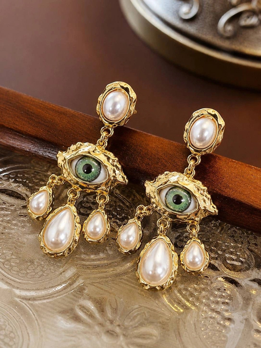 Close-up of gold evil eye pearl drop earrings resting on a carved metal tray showing textured detailing