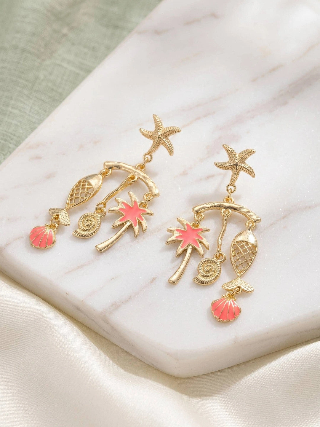 Pair of gold-plated tropical charm drop earrings with pink enamel shells and palm tree on marble surface