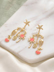 Pair of gold-plated tropical charm drop earrings with pink enamel shells and palm tree on marble surface