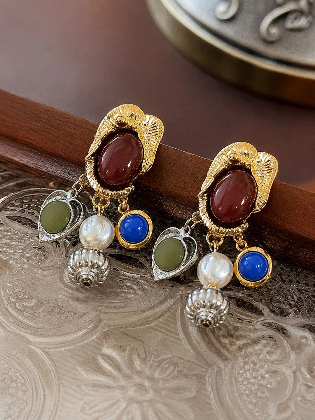 Close-up of two gold-tone drop earrings laid on a decorative silver tray showing red cabochon and mixed charms