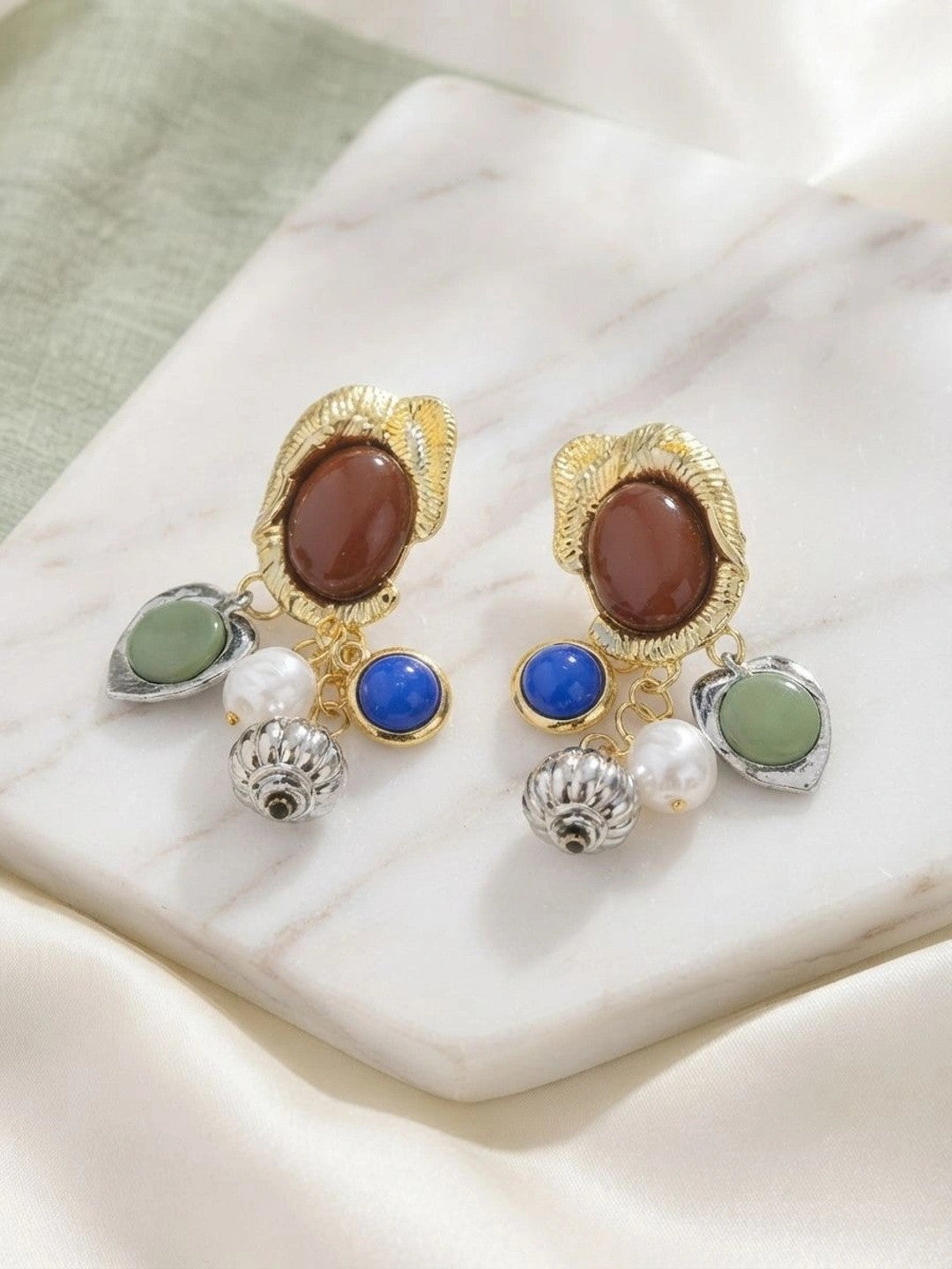 Pair of gold and silver-tone multi-charm drop earrings displayed on a white marble surface with green fabric backdrop