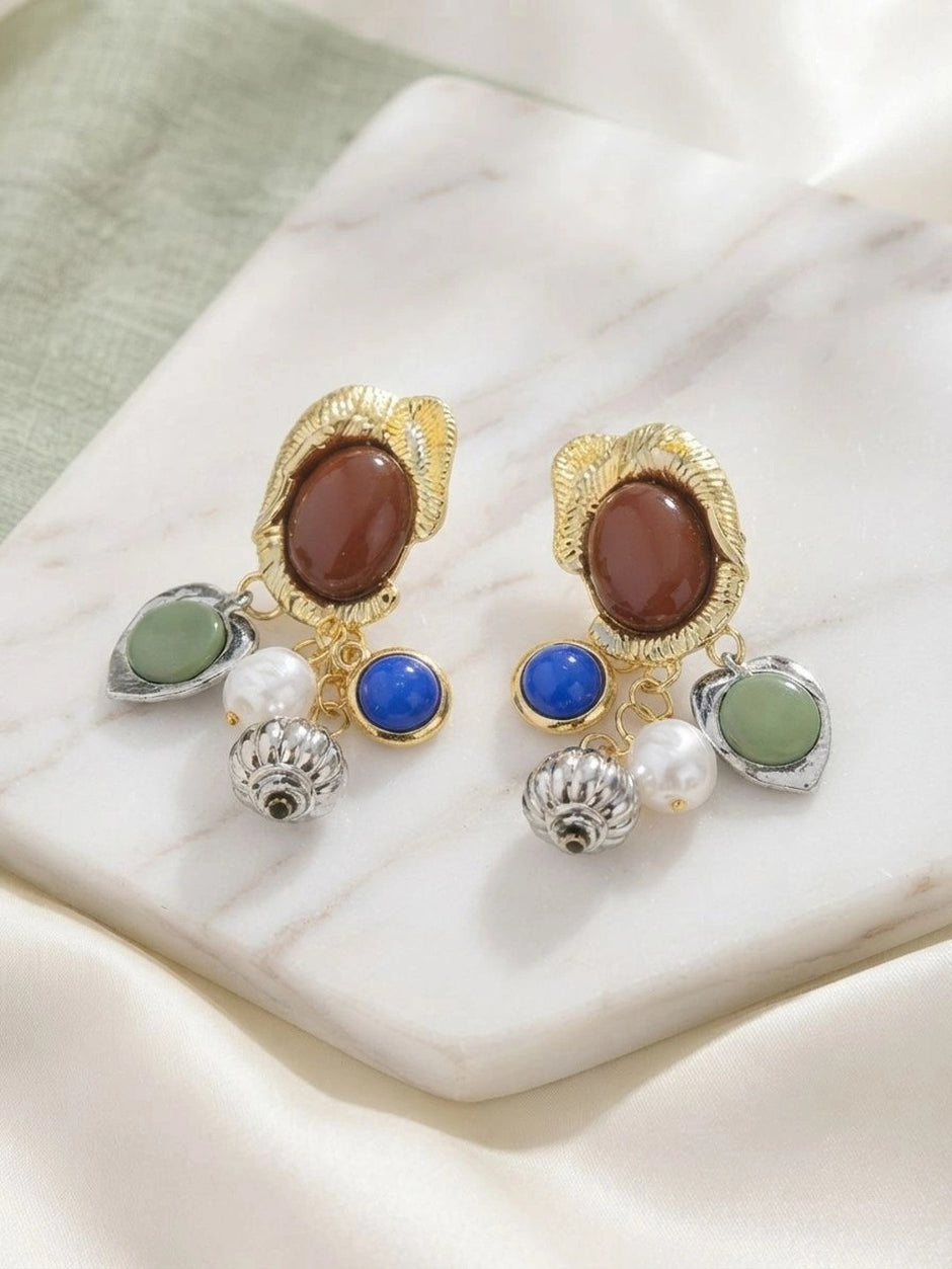 Pair of gold and silver-tone multi-charm drop earrings displayed on a white marble surface with green fabric backdrop